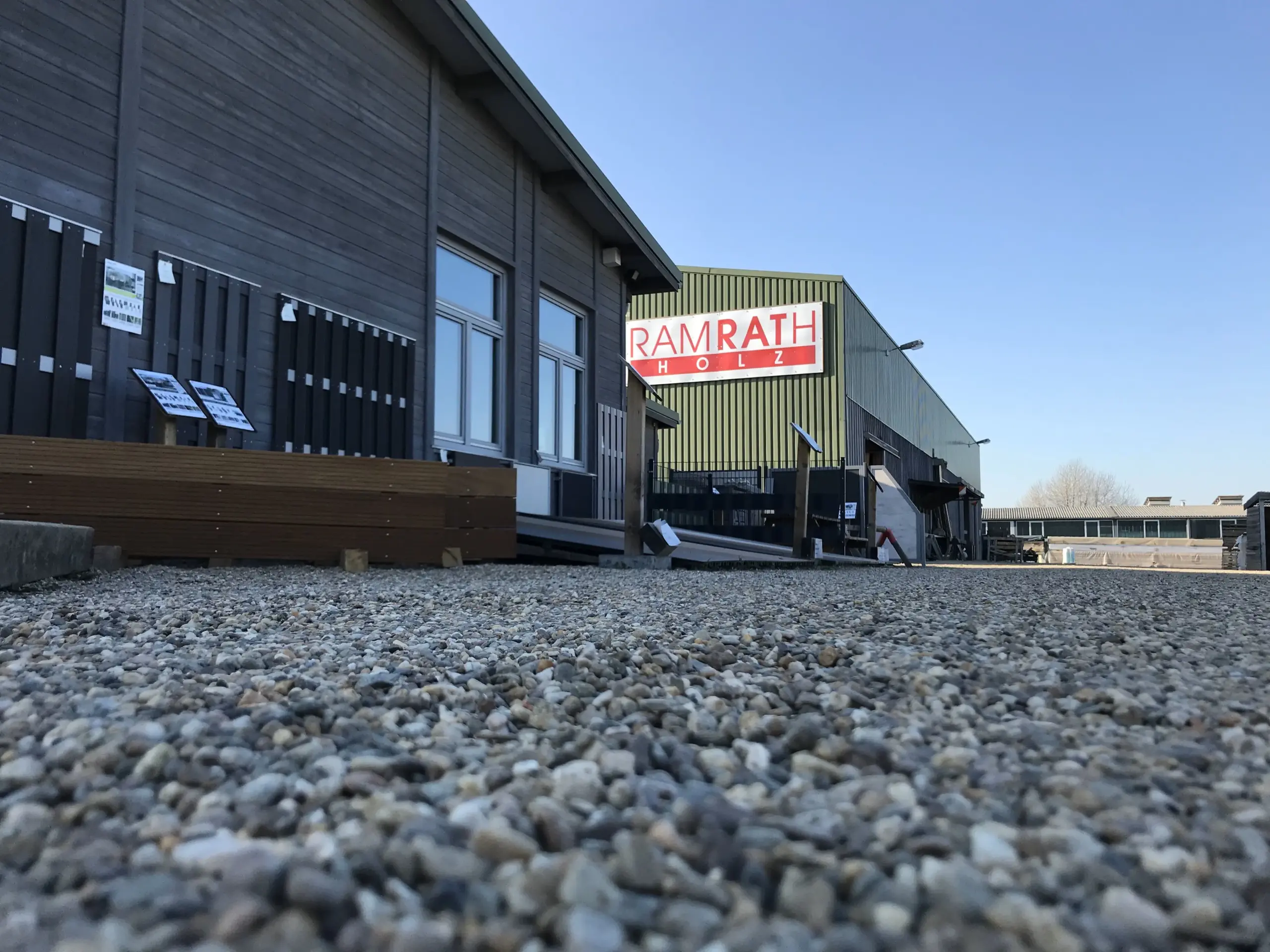 Ausstellungsgelände mit Zäunen, Terrassen und Lagerhalle – Blick von der Einfahrt Einfahrt mit Blick auf die Ausstellung für Zäune und Terrassen sowie die Lagerhalle im Hintergrund.